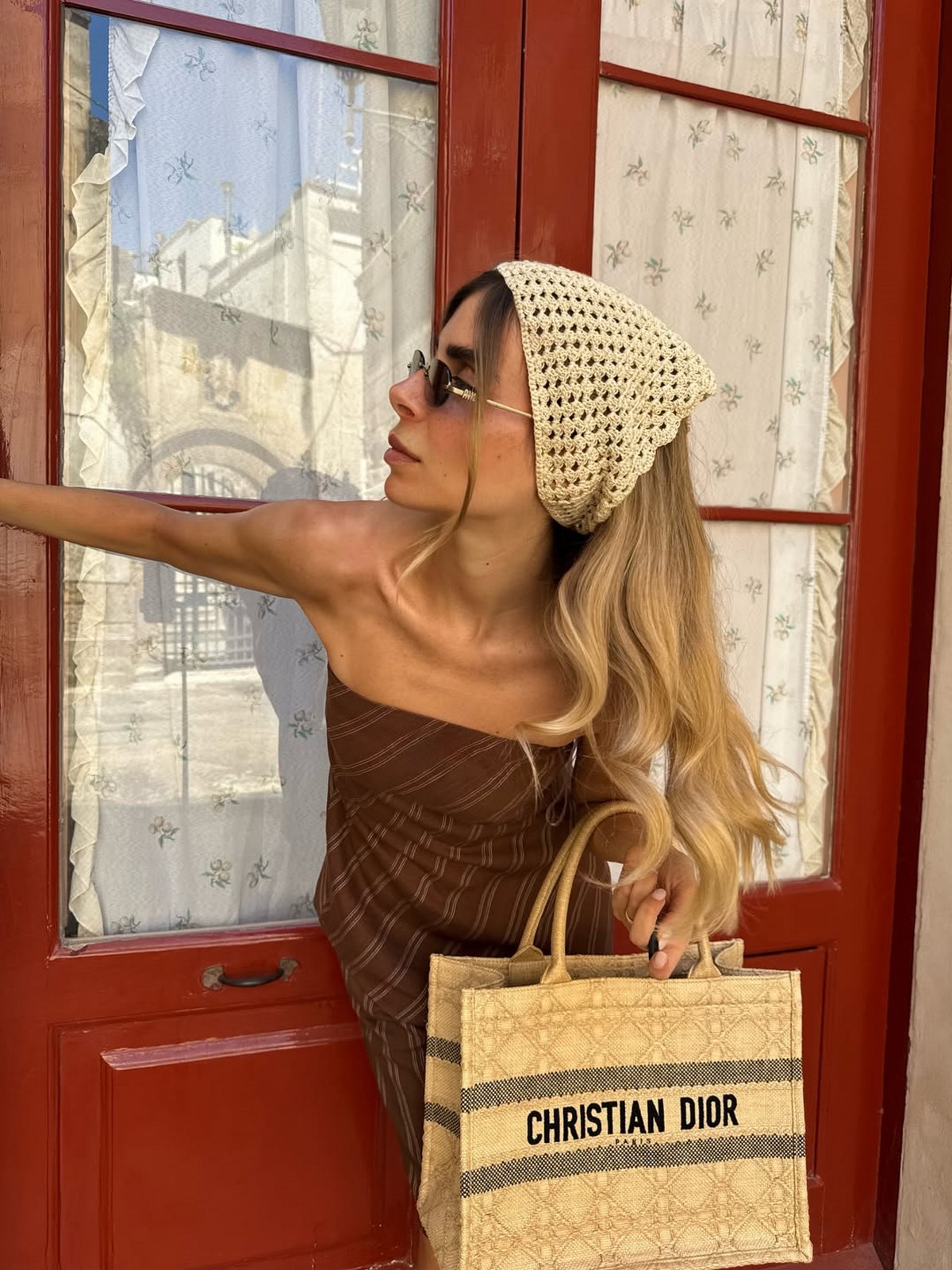 Женщина в кремовой вязаной косынке, коричневом топе и с бежевой сумкой у красной двери